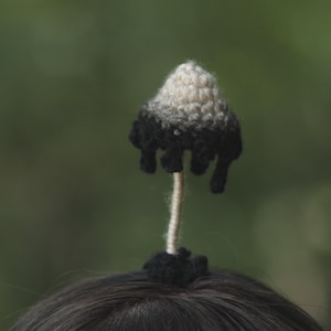 May include: A handcrafted crochet mushroom hair accessory. The mushroom cap is white and grey, fading to black at the edges. It's attached to a thin, beige stem and a black base, set against a blurred green backdrop.