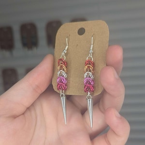 May include: A pair of handmade earrings with a chainmail design. The earrings feature a series of colorful rings in red, orange, gold, silver, and pink, with a silver spike pendant. The earrings are displayed on a brown card.