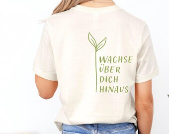 May include: Cream-colored t-shirt with a green plant graphic and the German text "WACHSE ÜBER DICH HINAUS". The shirt also features a "Cheeky Lemon Club" logo. The shirt is paired with blue jeans.