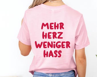 May include: A light pink t-shirt with the German text "MEHR HERZ WENIGER HASS" in red, meaning "More Heart, Less Hate." The shirt is worn by a person with blonde hair and blue jeans. The logo for "Cheeky Lemon Club" is in the upper right corner.