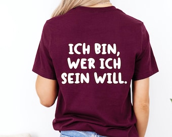 May include: A maroon t-shirt with the German phrase "ICH BIN, WER ICH SEIN WILL" printed in white. The shirt is a crew neck style and is being worn by a person with blonde hair.