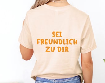 May include: A beige t-shirt with the German text "SEI FREUNDLICH ZU DIR" in orange letters. The shirt is being worn by a person with blonde hair and blue jeans. The logo for "Cheeky Lemon Club" is visible in the upper right corner.