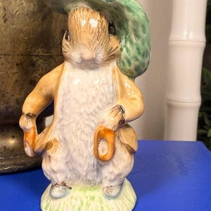 May include: A ceramic figurine of a rabbit wearing a green hat with an orange accent. The rabbit is dressed in a tan coat and white shirt, holding a small orange object. The figurine is standing on a green grassy base.