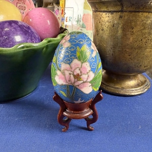 May include: A decorative egg with a blue background and a floral design in pink, white, and green, displayed on a small wooden stand. The egg is likely made of enamel or a similar material, and the stand is a dark brown color.
