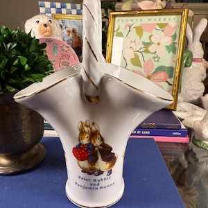 May include: White ceramic basket with a gold trim and handle. The basket features an illustration of Peter Rabbit and Benjamin Bunny holding a red basket of berries. The text "Peter Rabbit and Benjamin Bunny" is printed below the illustration.