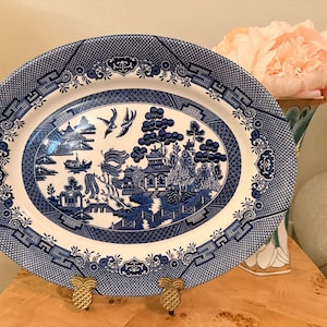 May include: Oval blue and white ceramic platter with a Willow pattern design. The platter features a detailed landscape scene with pagodas, trees, and birds. The rim has a decorative border. The platter is supported by two gold-colored pineapple-shaped stands.