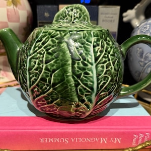 May include: A ceramic teapot shaped like a head of cabbage with a glossy green finish, featuring realistic embossed leaf textures, a curved spout, a sturdy handle, and a matching lid with a leaf-shaped finial, resting on a pink book.