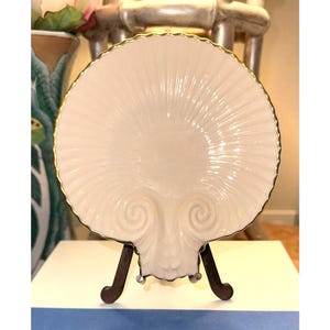 May include: A cream-colored, shell-shaped decorative plate with a gold trim, displayed on a dark brown metal stand. The plate features a detailed, ridged design radiating from the center, with two curled accents at the base.