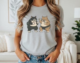 Puede incluir: Camiseta gris claro con una ilustración de dibujos animados de dos gatos luchadores de sumo. Un gato es blanco y negro, el otro es naranja y marrón. Ambos gatos visten atuendos de luchadores de sumo y tienen expresiones enfadadas.
