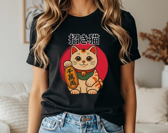May include: Black t-shirt with a cartoon Maneki-neko cat design. The cat is tan with orange stripes, a green collar, and holds a gold coin. Japanese text is above the cat, which is set against a red circle.