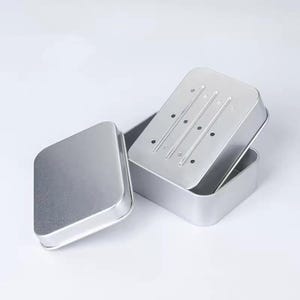 May include: A silver-colored rectangular soap dish with a lid. The lid has three parallel slots and several small holes for drainage. The dish is open, revealing the interior. The lid is slightly ajar, and another lid is to the left.