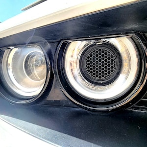 May include: Close-up of a car's headlight assembly. The assembly features a bright, circular headlight with a chrome trim and a black honeycomb-patterned center. The surrounding bodywork is white and black.