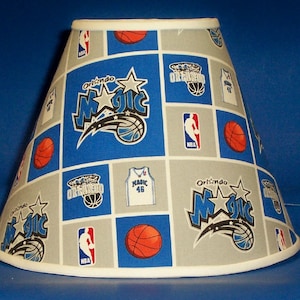 May include: A white lampshade with a blue and gray pattern featuring the Orlando Magic basketball team logo, basketballs, and the NBA logo.