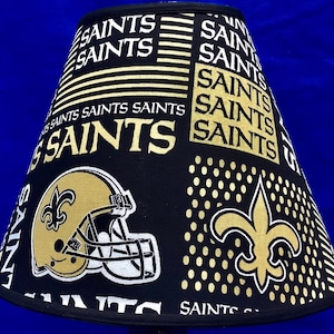 May include: A black and gold lampshade with a Saints logo and repeated text "Saints".