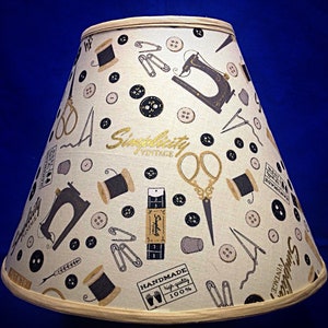 May include: A beige lampshade with a gold trim features a repeating pattern of sewing-themed illustrations, including buttons, spools of thread, scissors, sewing machines, and safety pins. The lampshade also features the text "Simplicity Vintage" and "Handmade high quality 100%".