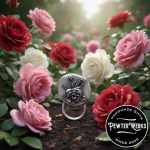 May include: A silver-toned metal ring featuring a circular face embossed with a detailed rose and leaf design. The ring is positioned among red, pink, and white roses. A black circular logo in the corner reads HANDMADE GIFTS PEWTER WERKS SINCE 2025.
