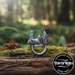 May include: A silver-toned metal ring featuring a sculptural design of two pine trees flanking a small mountain peak. The band is circular and smooth. A circular black logo in the corner displays the text: HANDMADE GIFTS PEWTER WERKS SINCE 2025.