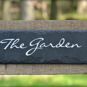 May include: A black slate sign with white lettering that reads "The Garden". The sign is mounted on a wooden fence.