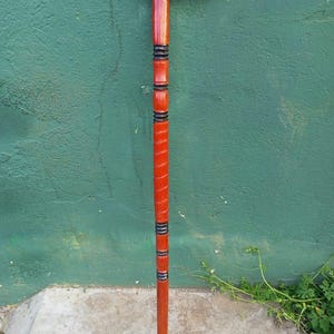 May include: A red wooden walking cane with a curved handle. The cane has a series of black and red bands along the shaft. The cane is standing upright against a green wall.