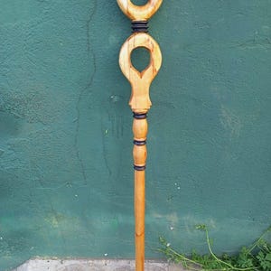 May include: A handcrafted wooden walking stick with a unique design. The top features a circular handle and a decorative cutout. The shaft is long and slender, with black accents. The wood has a natural, light brown finish.