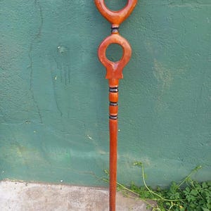 May include: A wooden walking stick with a dark brown finish. The top features a large, rounded loop above a smaller, similar loop. The shaft is long and slender, with black and brown accents near the top. The bottom has a black tip.