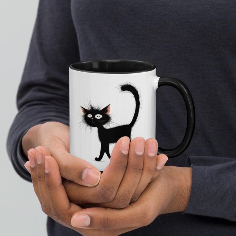 May include: A white ceramic mug with a black interior and handle. The mug features a cartoon illustration of a black cat with wide eyes and a fluffy tail. The cat is walking on a white surface.