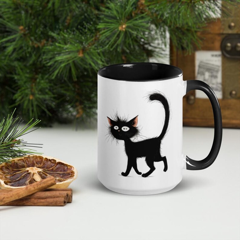 May include: White ceramic mug with a black interior and handle. The mug features a cartoon illustration of a black cat with wide eyes and a fluffy tail. The mug is set against a background of greenery and a wooden chest.