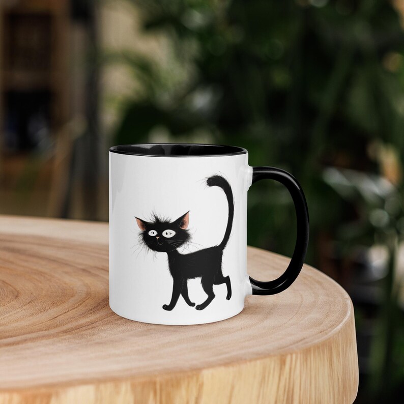 May include: White ceramic mug with a black interior and handle. The mug features a cartoon illustration of a black cat with wide eyes and a curved tail. The cat is walking on a white background.