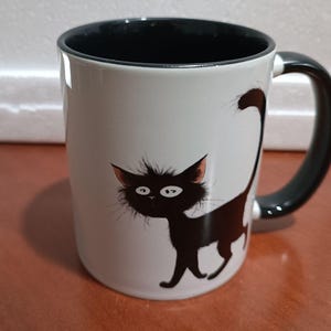 May include: White ceramic mug with a black interior and handle. The mug features a whimsical illustration of a black cat with large eyes and a fluffy tail. The cat is walking on the side of the mug.