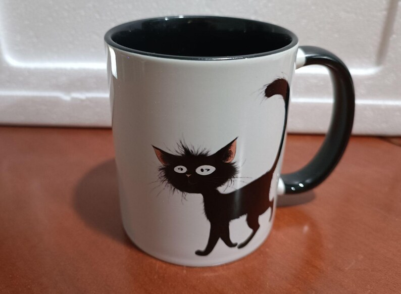 May include: White ceramic mug with a black interior and handle. The mug features a whimsical illustration of a black cat with large eyes and a fluffy tail. The cat is walking on the side of the mug.