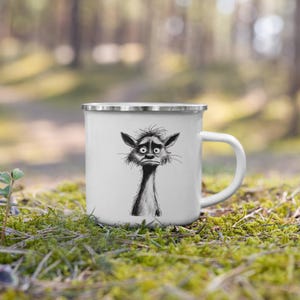 May include: White enamel mug with a silver rim and handle. The mug features a black and white illustration of a creature with a worried expression. The mug is set against a blurred background of green foliage.