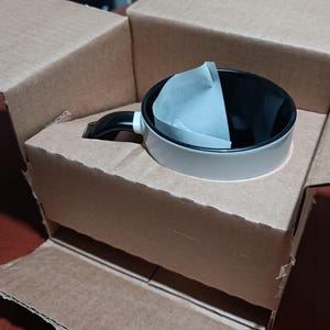 May include: A white and black coffee mug with a black handle and a paper filter inside, packaged in a brown cardboard box. The mug has a light gray exterior and a black interior. The box is open, revealing the mug nestled inside.