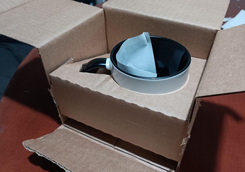 May include: A white and black coffee mug with a black handle and a paper filter inside, packaged in a brown cardboard box. The mug has a light gray exterior and a black interior. The box is open, revealing the mug nestled inside.
