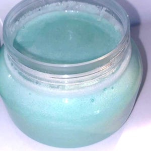 May include: A clear glass jar filled with a light blue, creamy substance. The jar has a transparent lid and sits on a white surface. The contents appear smooth and slightly foamy, suggesting a cosmetic product.