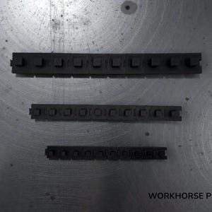 May include: Three black, rectangular, segmented components with square-shaped protrusions are arranged on a metallic surface. The components vary in size, with the largest at the top and the smallest at the bottom. The text "WORKHORSE PRINTS" is visible at the bottom right.