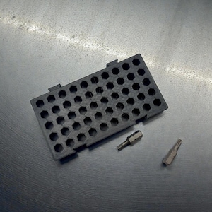 May include: A black, rectangular tool organizer with numerous hexagonal holes, accompanied by two silver-colored bits. The organizer is designed to hold small tools, and is set against a textured, metallic surface.