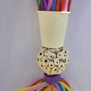May include: A colorful bird toy featuring a cream-colored cup filled with assorted straws in shades of purple, blue, pink, and orange. Below the cup is a textured sphere, and below that, a purple cap with colorful pipe cleaners extending outwards. A fun, engaging toy.
