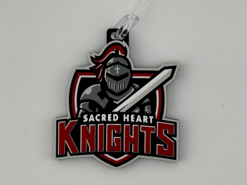 May include: A rectangular tag featuring the words "SACRED HEART KNIGHTS" in red letters. The tag has a black border and a shield design with a knight in gray armor holding a sword. A clear plastic loop is attached.