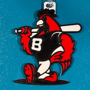 May include: A red and black baseball-themed ornament featuring a cartoon cardinal holding a baseball bat. The cardinal wears a black jersey with a white "B" on the front. The ornament has a white loop for hanging.