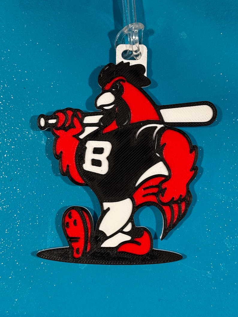 May include: A red and black baseball-themed ornament featuring a cartoon cardinal holding a baseball bat. The cardinal wears a black jersey with a white "B" on the front. The ornament has a white loop for hanging.