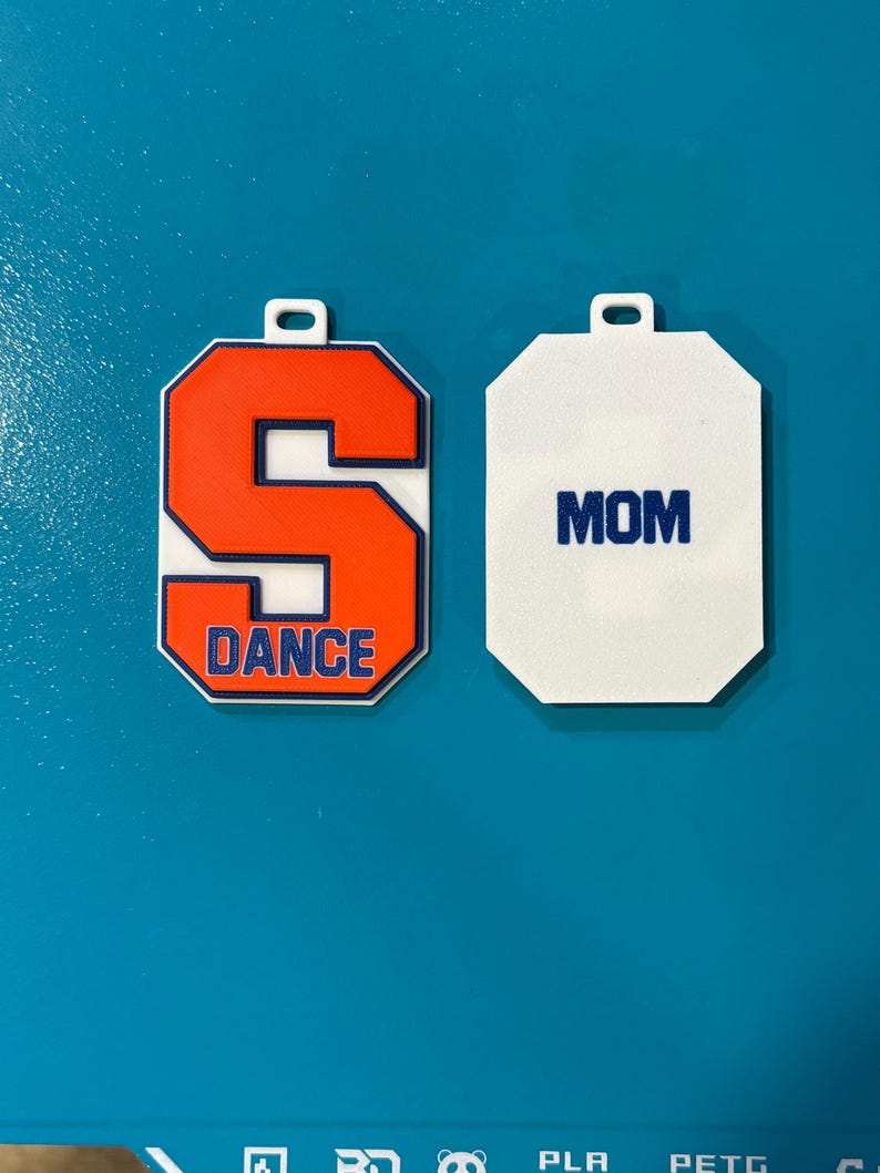 May include: Two white and orange keychains. One keychain features the letter "S" in orange with a blue outline and the word "DANCE" in blue. The other keychain is white with the word "MOM" in blue.