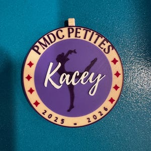 May include: A round, multi-layered ornament with the text "PMDC PETITES" in a cream-colored font. The center features a purple circle with a ballerina silhouette and the name "Kacey" in white script. Red star accents and the years 2025-2026 are also present.