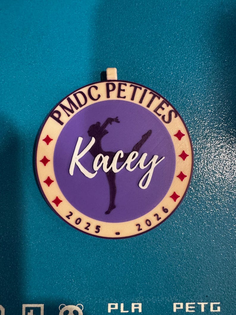 May include: A round, multi-layered ornament with the text "PMDC PETITES" in a cream-colored font. The center features a purple circle with a ballerina silhouette and the name "Kacey" in white script. Red star accents and the years 2025-2026 are also present.
