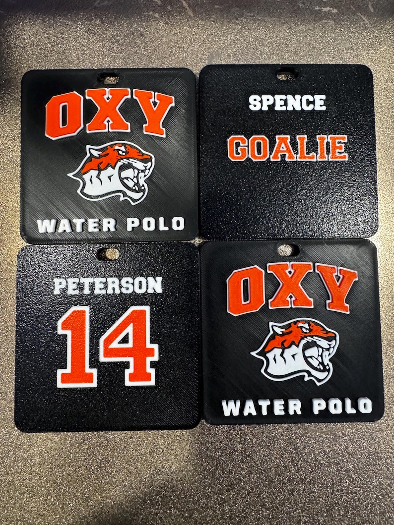May include: Four black square tags with white and orange text. Two tags read "OXY" with a tiger logo and "WATER POLO". One tag says "SPENCE GOALIE". The last tag says "PETERSON 14". Each tag has a small hole.