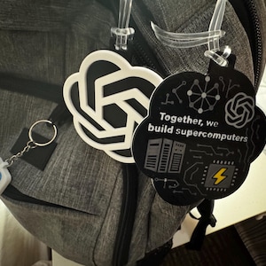 May include: A gray backpack with a keychain and two cloud-shaped tags. One tag is white with a black design, and the other is black with the text "Together, we build supercomputers" and a yellow lightning bolt.