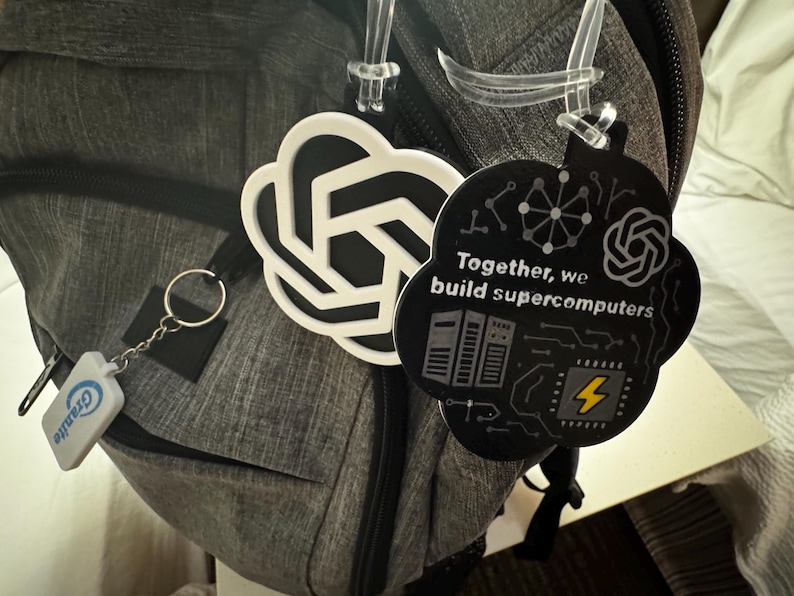 May include: A gray backpack with a keychain and two cloud-shaped tags. One tag is white with a black design, and the other is black with the text "Together, we build supercomputers" and a yellow lightning bolt.