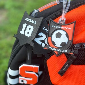 May include: Black and orange soccer-themed bag tags. One tag displays "MICHALS" and the number "18". Another tag shows "LA" and the number "2". A third tag features a soccer ball graphic and the text "STOUGHTON YOUTH SOCCER".