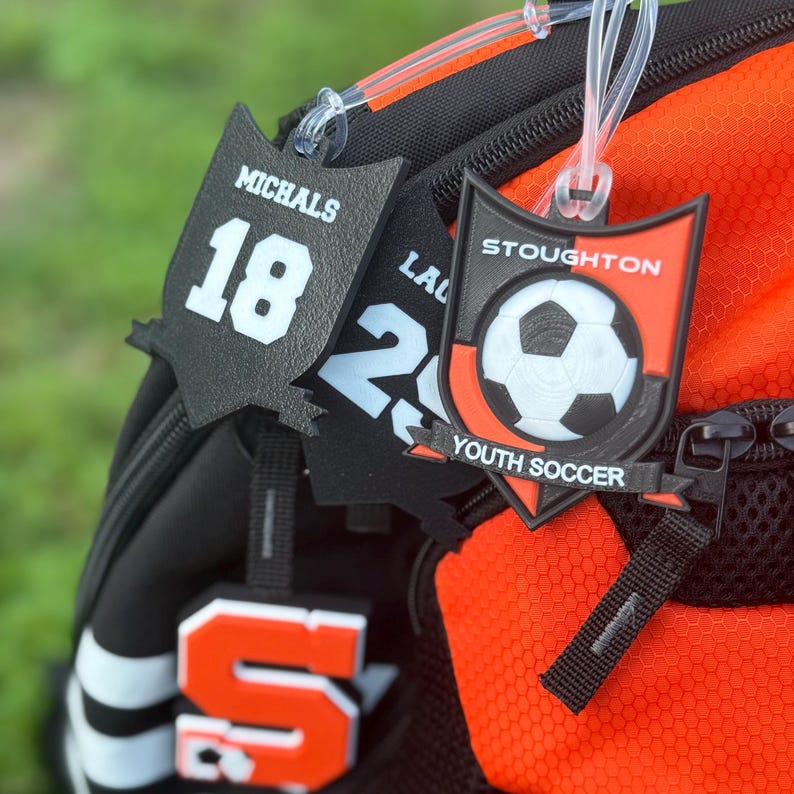 May include: Black and orange soccer-themed bag tags. One tag displays "MICHALS" and the number "18". Another tag shows "LA" and the number "2". A third tag features a soccer ball graphic and the text "STOUGHTON YOUTH SOCCER".
