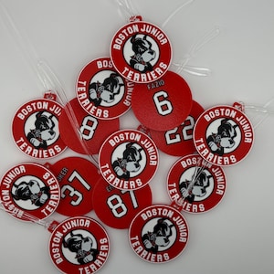 May include: A collection of red and white Boston Junior Terriers luggage tags. Each tag features a bulldog mascot and the words "Boston Junior Terriers." Some tags display numbers and the name "FAZIO."