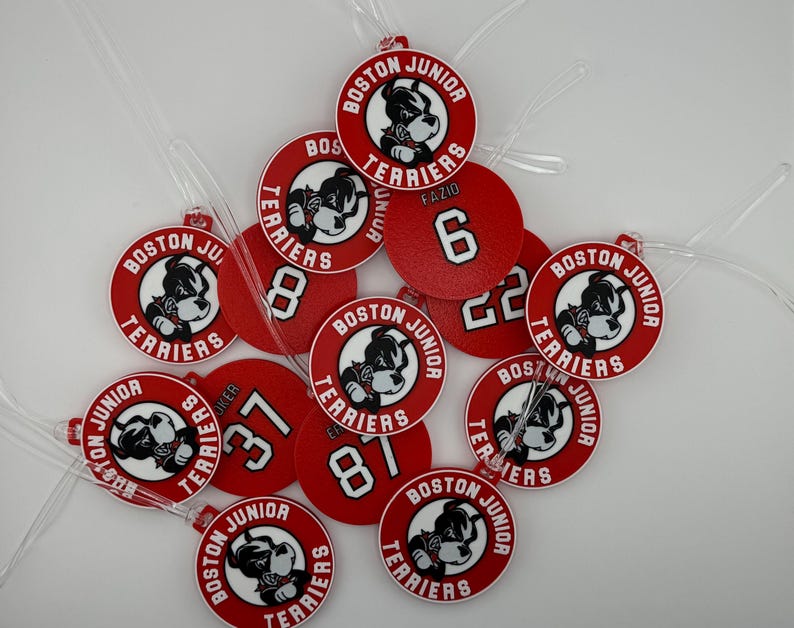 May include: A collection of red and white Boston Junior Terriers luggage tags. Each tag features a bulldog mascot and the words "Boston Junior Terriers." Some tags display numbers and the name "FAZIO."